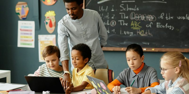 Children Using Laptops at Desk while Male Teacher Helping Kids in Classroom