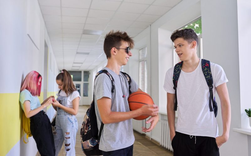 Group of teenage students talking standing outside school building, two males with backpacks basketball ball in focus, schoolgirls in corridor background. Lifestyle, communication, youth concept