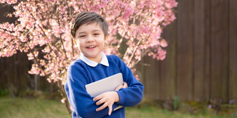 Portrait happy Kid holding tablet pc waiting for School bus in morning, Positive young boy with school uniform looking at camera while standing in the garden. Education and Back to school concept