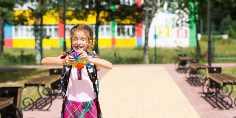 Little girl with a backpack and in a school uniform in the school yard plays pop it toy. Back to school, September 1. The pupil relaxes after lessons. Primary education, elementary class for student