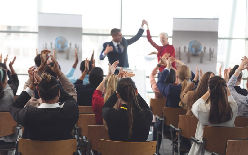 Rear view of diverse business people applauding and celebrating while they are sitting in front of multi-ethnic business executives at business seminar in office building